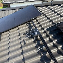 Solcity - Fotowoltaika i Systemy Grzewcze - Montaż paneli fotowoltaicznych na dachu z ciemnej dachówki. Widoczne elementy mocujące i okablowanie. Częściowo zamontowane panele w górnej części kadru.