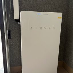 Solcity - Fotowoltaika i Systemy Grzewcze - Biały, matowy magazyn energii ATMOCE z niebieską diodą LED, umieszczony w rogu pomieszczenia z szarą, chropowatą ścianą i brązową podłogą.