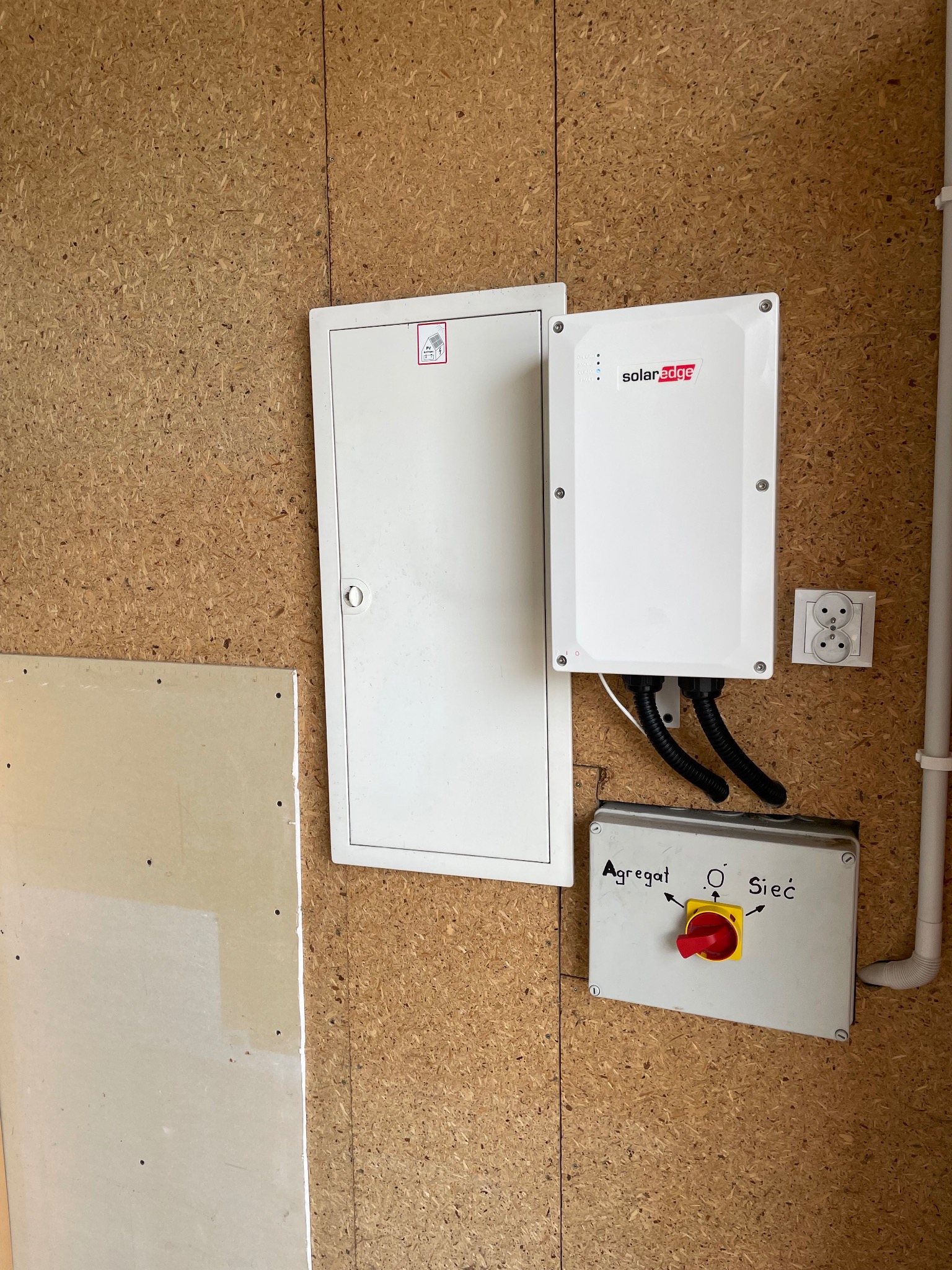 Biały inwerter SolarEdge zamontowany na płycie OSB, obok skrzynka z przełącznikiem agregat/sieć, gniazdko elektryczne i skrzynka rozdzielcza.