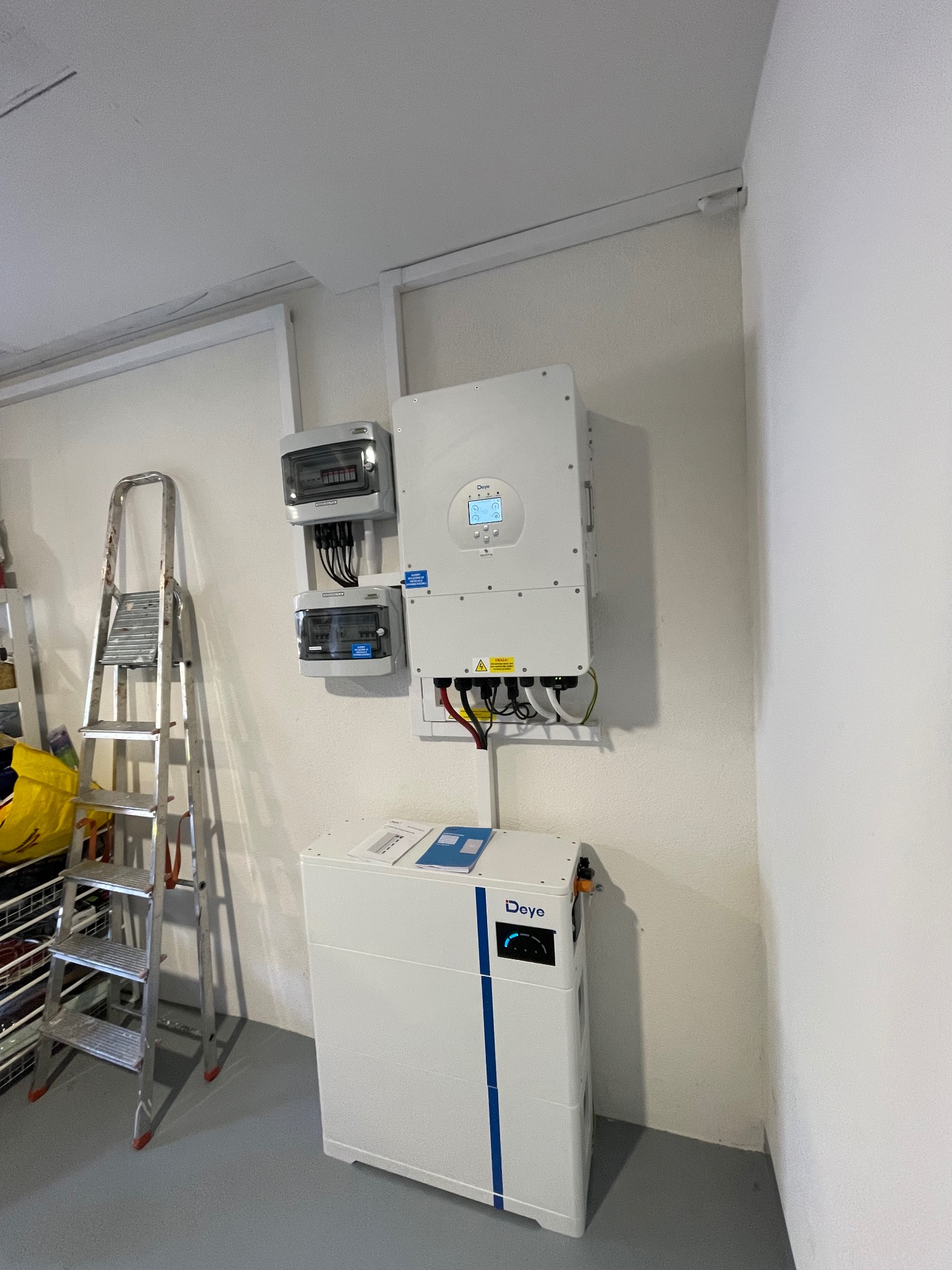 Biały magazyn energii Deye z niebieskimi paskami, falownik na ścianie, skrzynki elektryczne i aluminiowa drabina w tle, szara podłoga.