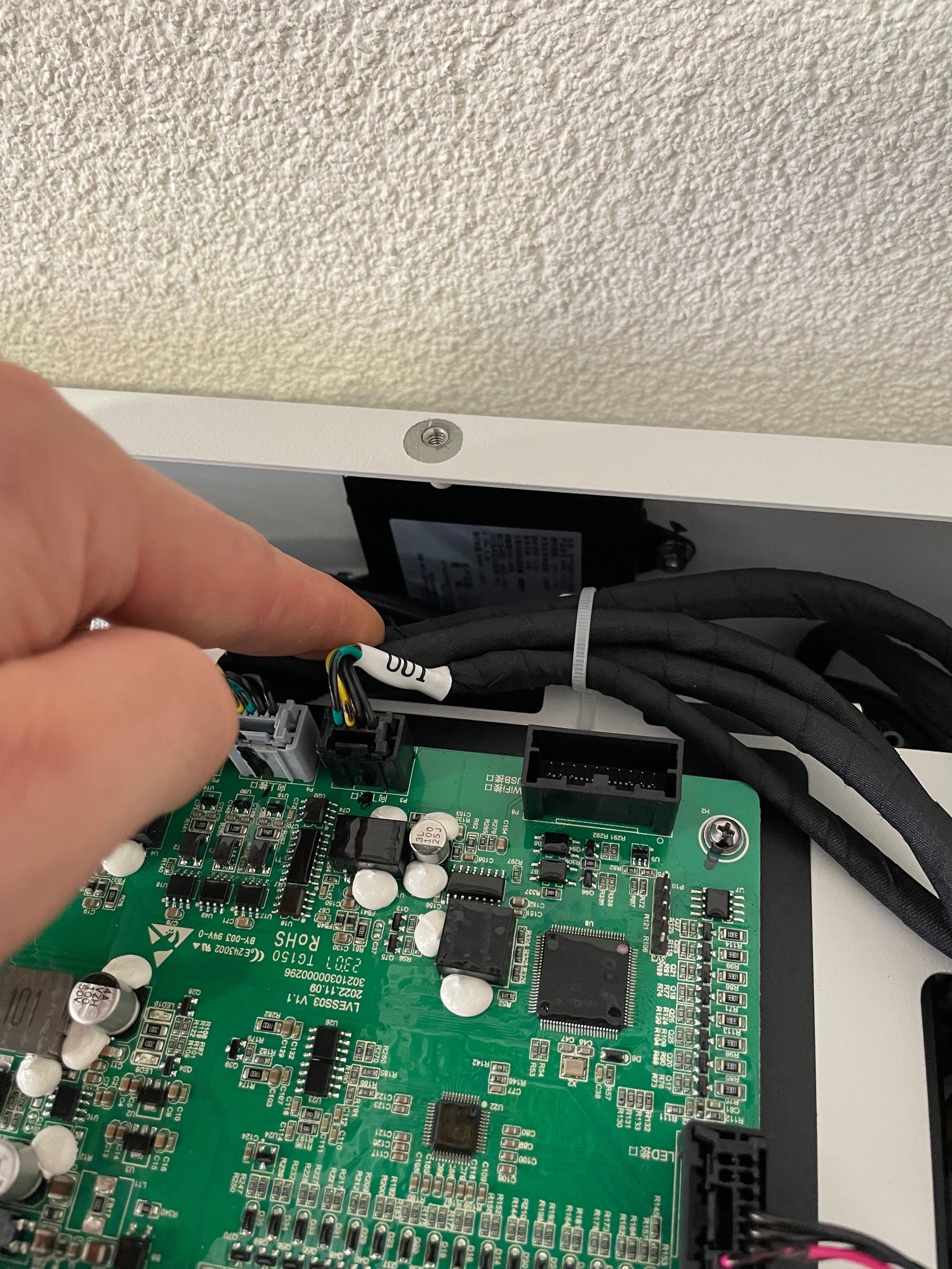 Szczegółowe ujęcie zielonej płytki drukowanej z elektroniką wewnątrz białej obudowy, z palcem wskazującym na złącze kablowe z czarno-żółto-zielonymi przewodami.