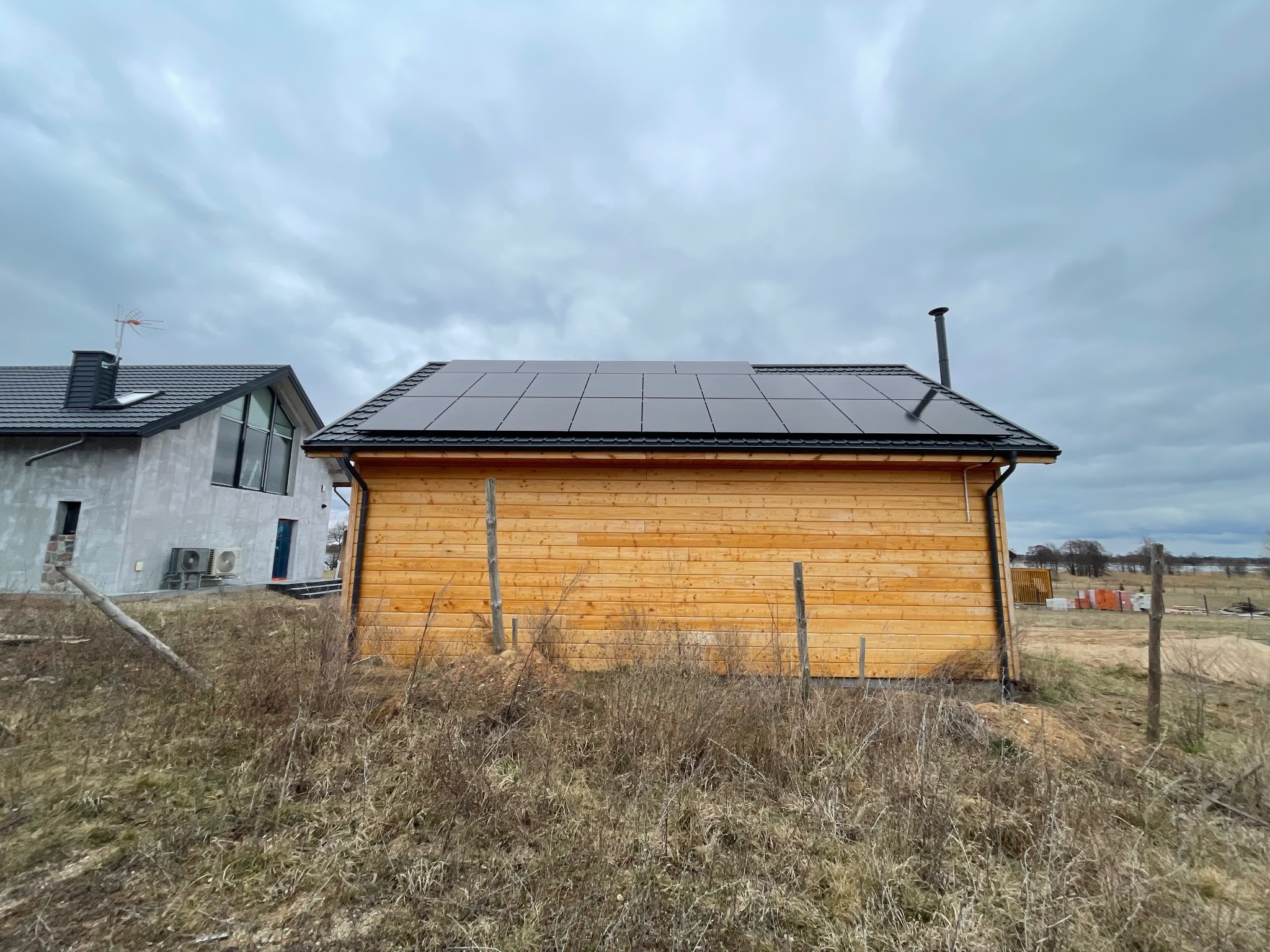 Drewniany dom z panelami słonecznymi na dachu, widok z poziomu gruntu na tle pochmurnego nieba i sąsiedniego budynku w stanie surowym.