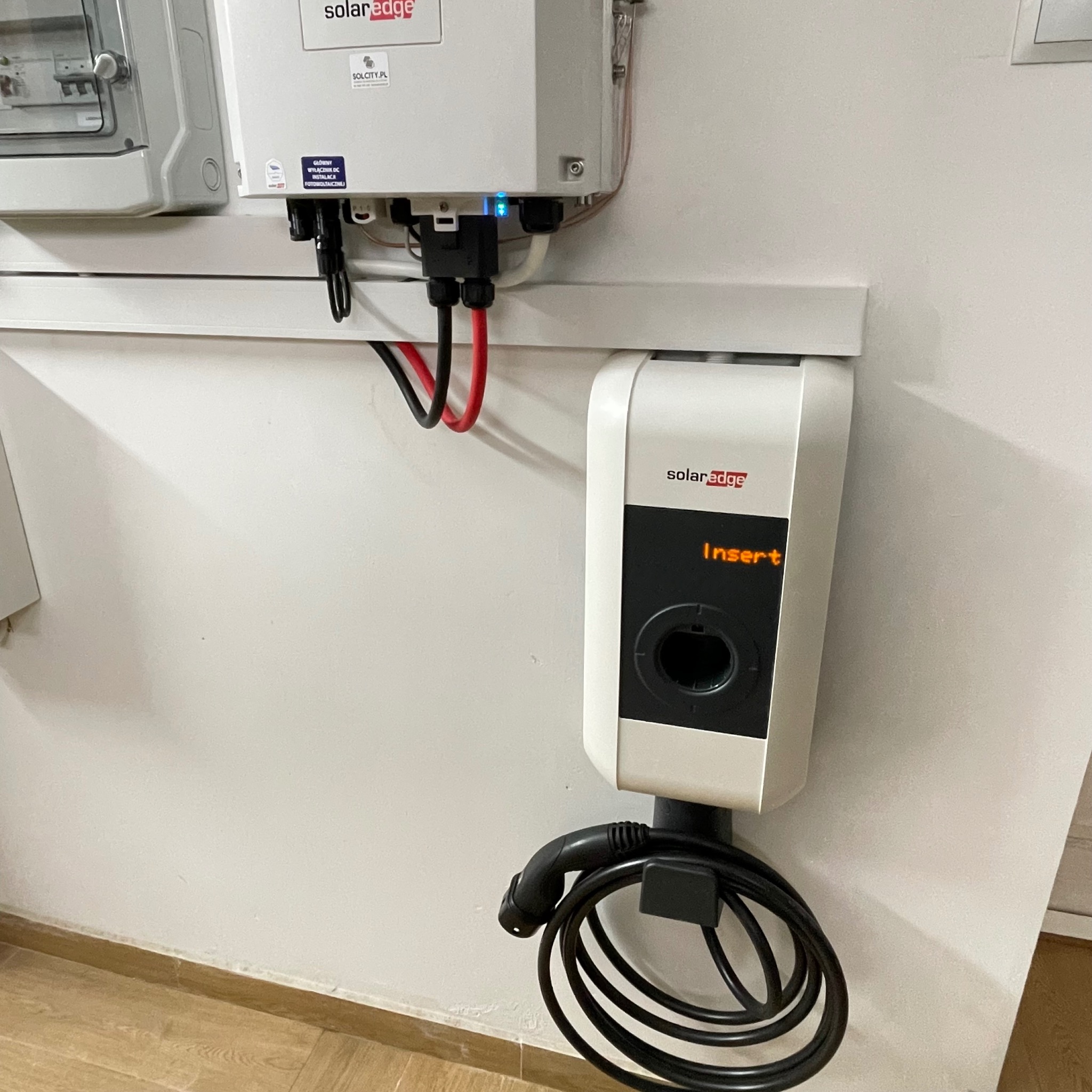 Biała stacja ładowania z napisem 'Insert' na ścianie, z podłączonym kablem. Obok widoczny inwerter z przewodami.