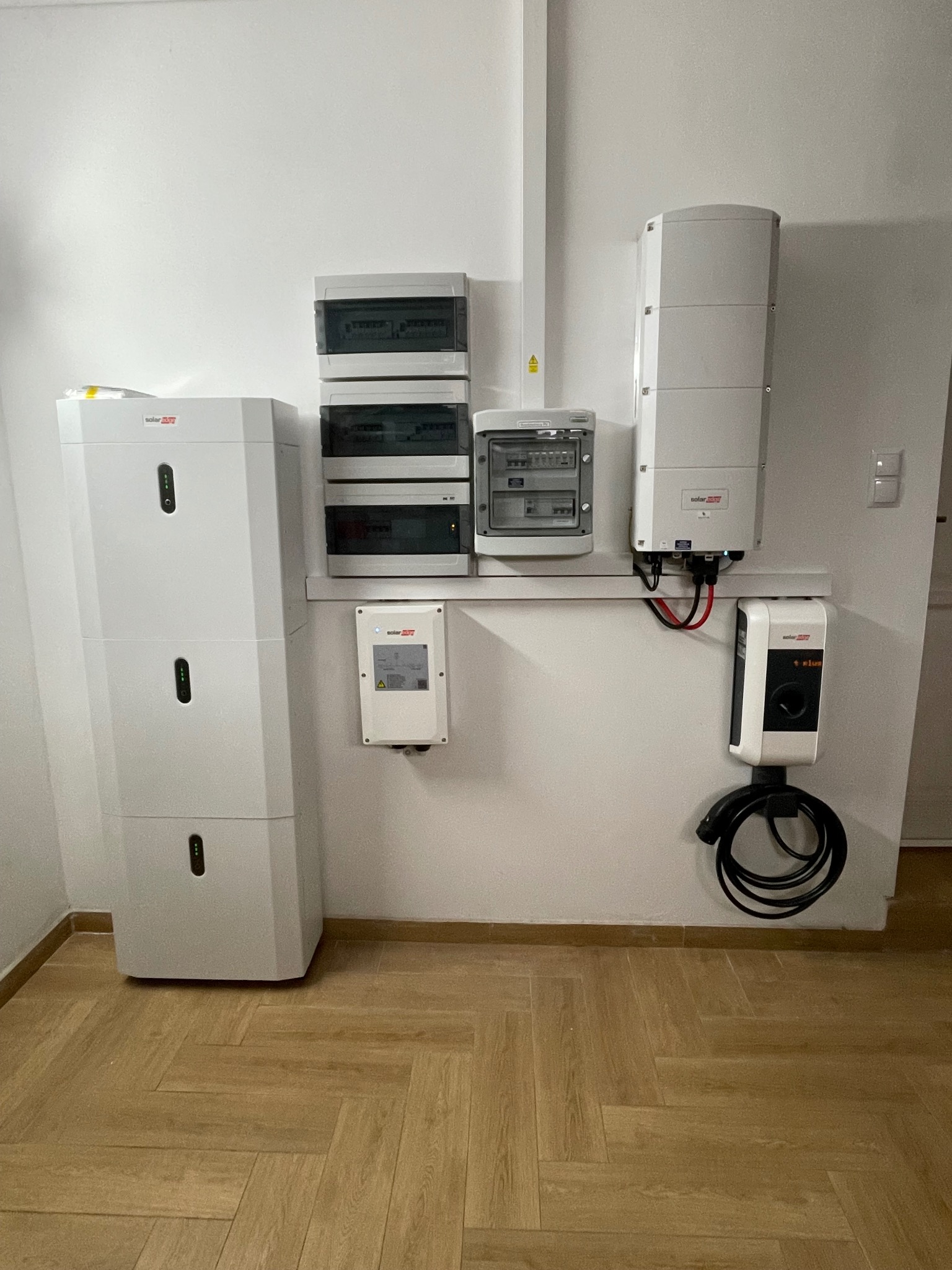 Wewnętrzna instalacja elektryczna z białymi skrzynkami rozdzielczymi, modułowym magazynem energii i ładowarką do pojazdów elektrycznych zamontowanymi na ścianie w pomieszczeniu z parkietem w jodełkę.