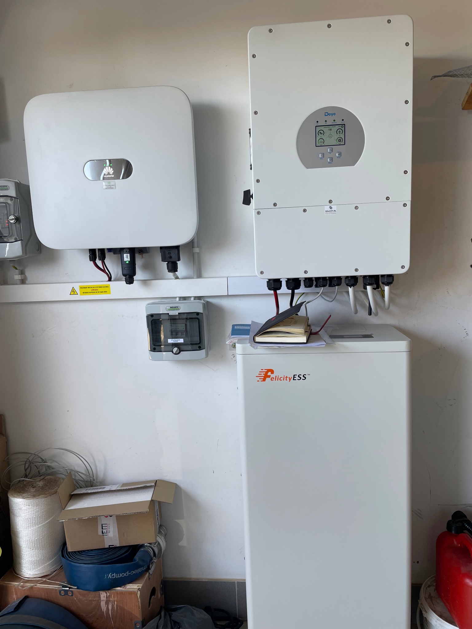 Dwa białe inwertery fotowoltaiczne marki Huawei i Daye oraz biały magazyn energii Felicity ESS zamontowane na ścianie, widoczne okablowanie i elementy instalacji elektrycznej.
