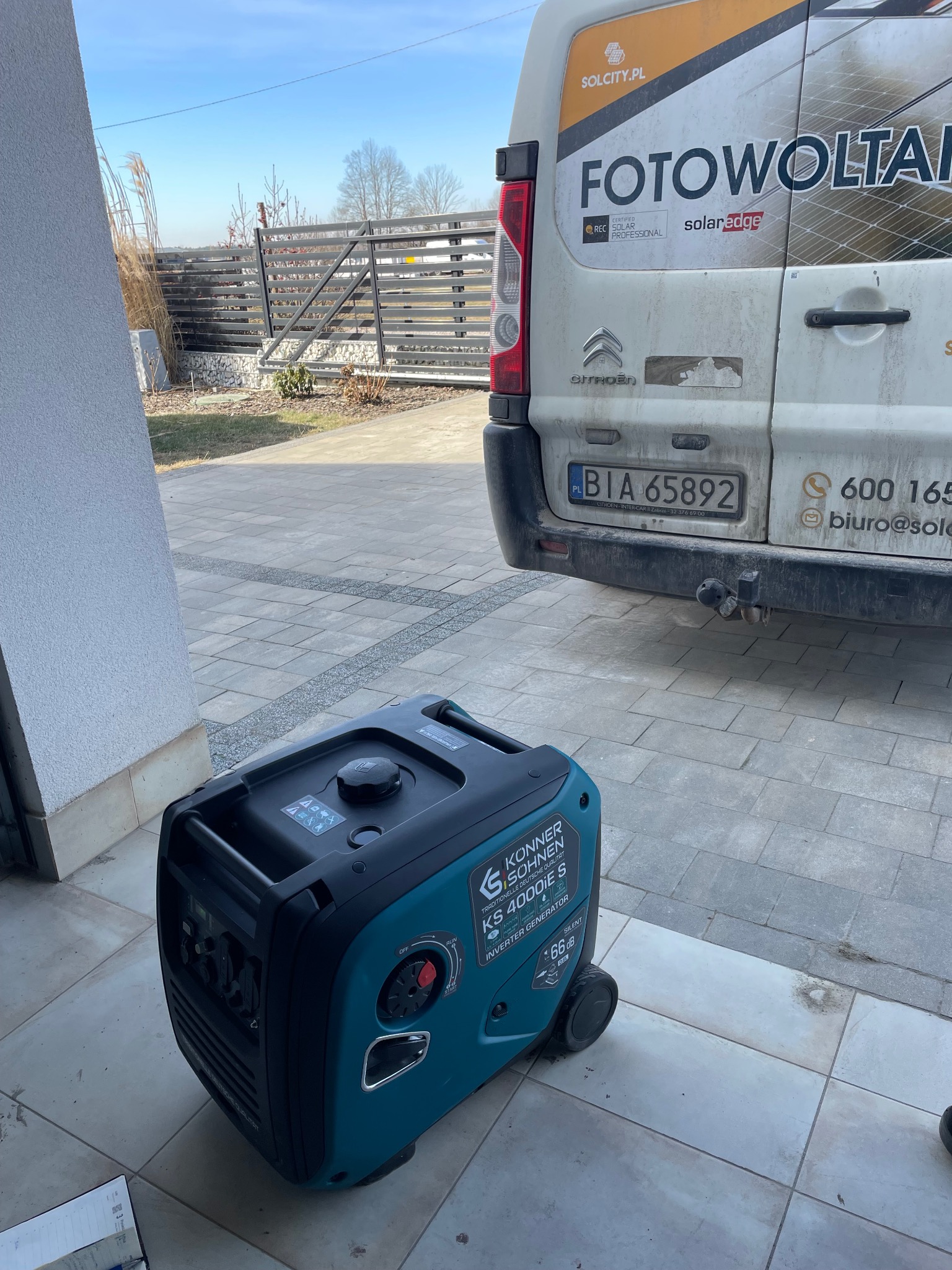 Agregat prądotwórczy inwertorowy Könner & Söhnen KS 4000iE S na tle zaparkowanego, lekko zabrudzonego samochodu dostawczego z reklamą firmy oferującej instalacje OZE.