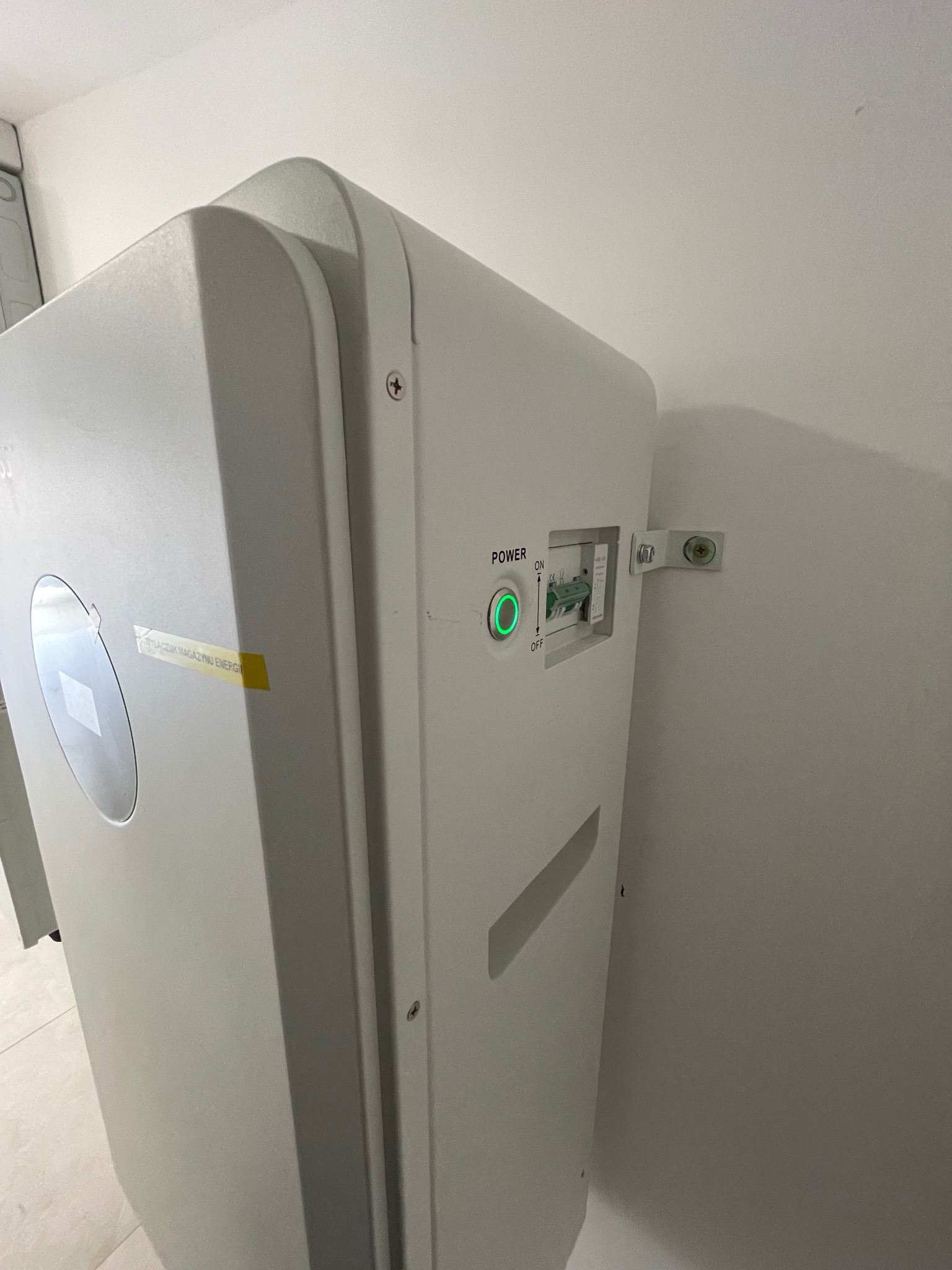 Biały moduł z włącznikiem zasilania w pozycji ON, przymocowany do białej ściany za pomocą metalowych wsporników.