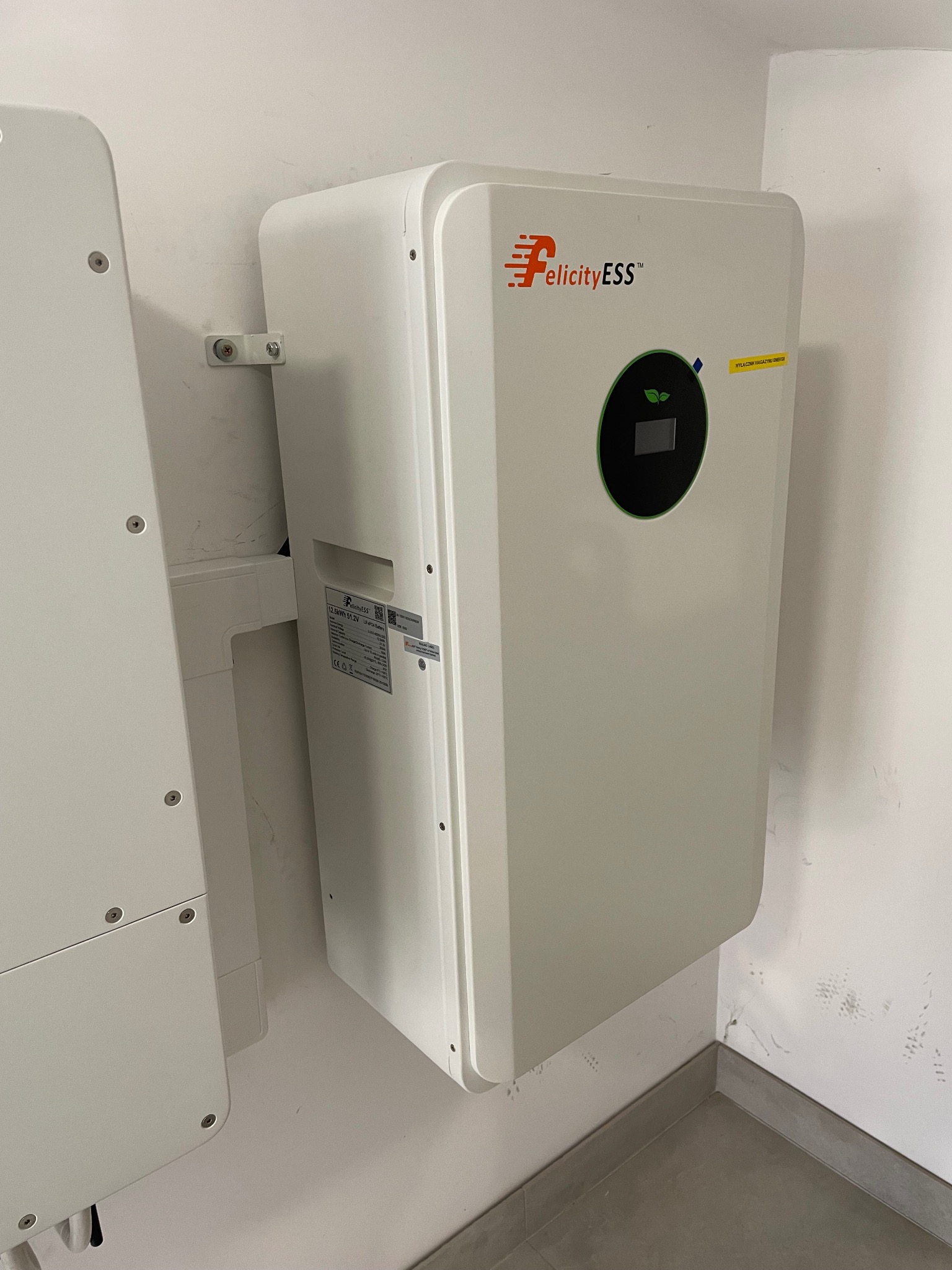 Biały magazyn energii Felicity ESS zamontowany na ścianie w Kurianach, widoczny fragment drugiego urządzenia po lewej stronie.