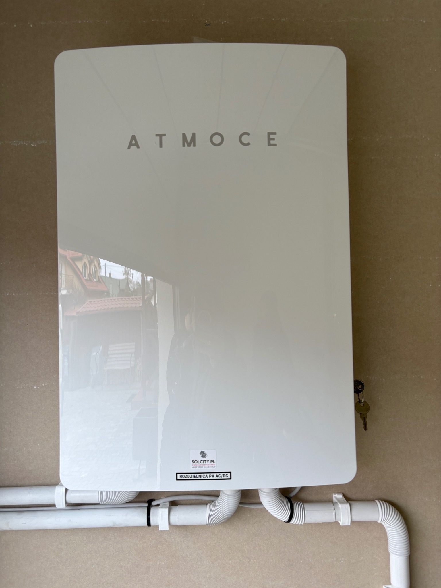 Biała rozdzielnica PV AC/DC marki ATMO CE zamontowana na ścianie, z widocznymi przewodami i kluczykiem. W tle odbicie budynku.
