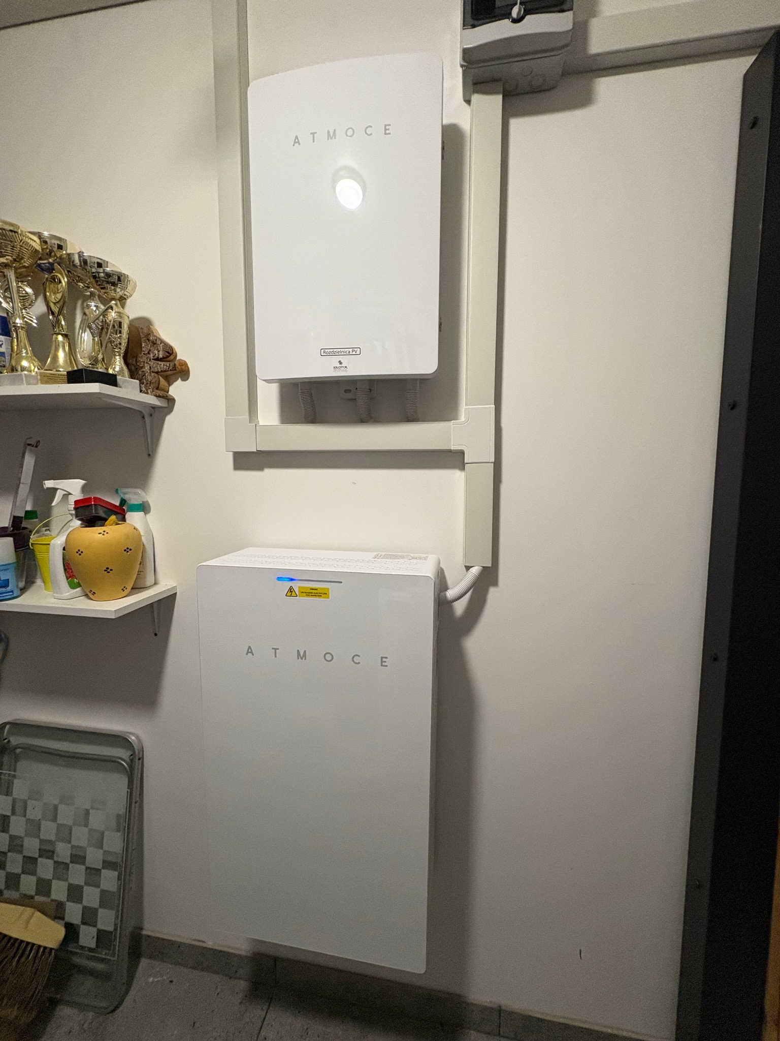 Dwa białe moduły ATMOCE zamontowane na ścianie w garażu, połączone listwami. Górny moduł z okrągłym wskaźnikiem, dolny z niebieską diodą. Obok półki z trofeami i środkami czystości.