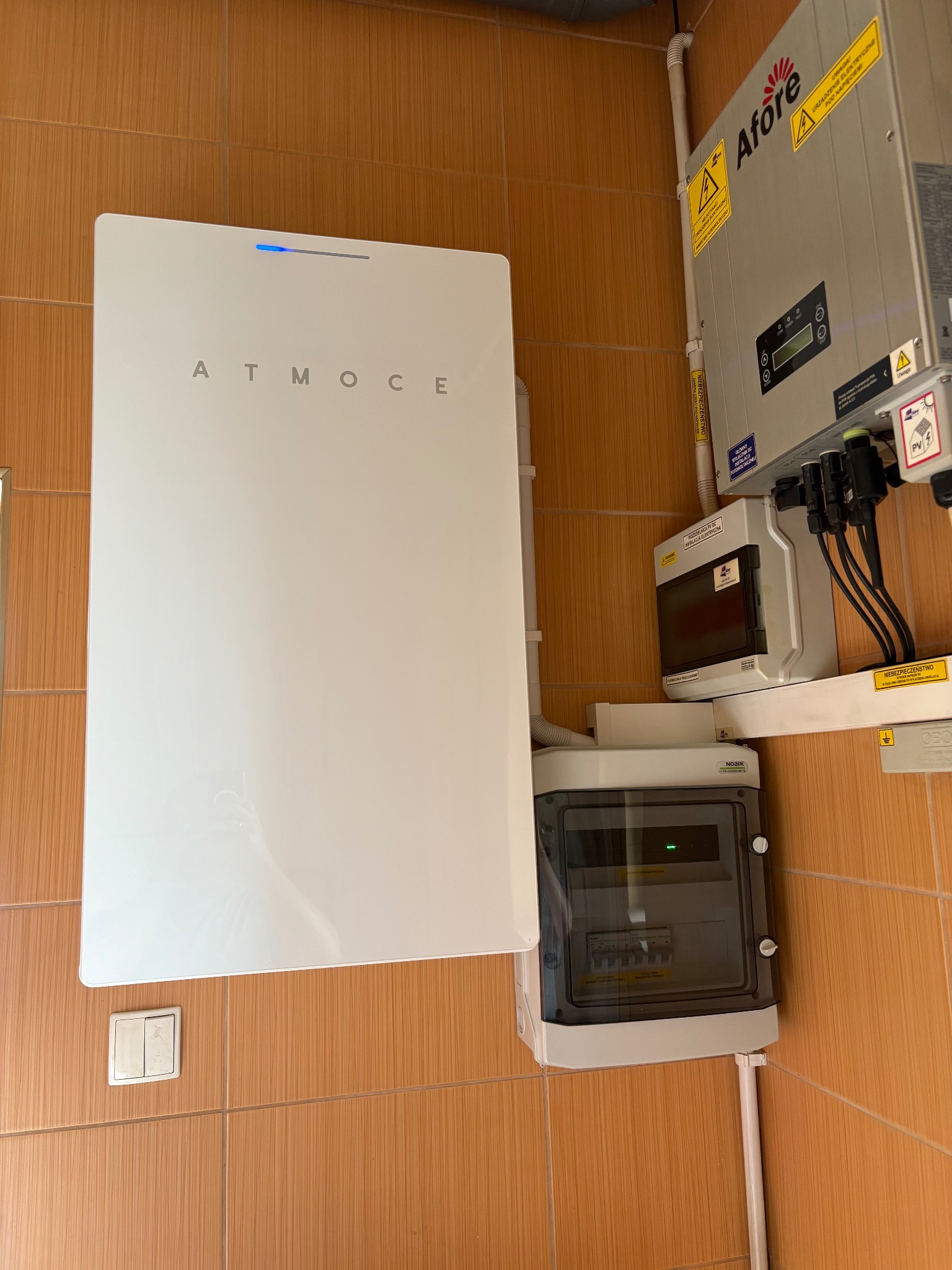 Biały akumulator ATMOCE z niebieską diodą, obok szary inwerter Afore i rozdzielnica elektryczna na brązowych płytkach. Elementy instalacji fotowoltaicznej w pomieszczeniu.