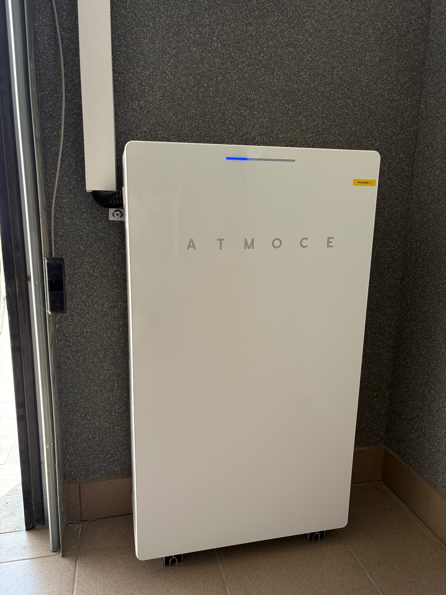 Biały, matowy magazyn energii ATMOCE z niebieską diodą LED, umieszczony w rogu pomieszczenia z szarą, chropowatą ścianą i brązową podłogą.