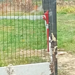 Czerwony hydrant przeciwpożarowy zamontowany przy metalowym ogrodzeniu panelowym z betonową podmurówką, widoczny dym wydobywający się z hydrantu.