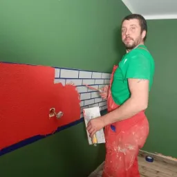 Mężczyzna w czerwonym kombinezonie i zielonej koszulce nakłada czerwoną gładź na ścianę z fragmentem imitacji białych cegiełek z szarą fugą, na tle zielonej ściany i drewnianej podłogi.