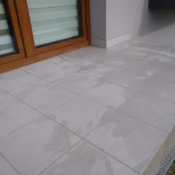 Wykończony taras z jasnymi, kwadratowymi płytkami ceramicznymi, widoczne ślady po myciu, drewniana rama okienna, jasna elewacja budynku.