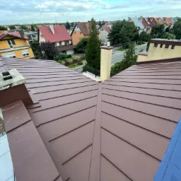 Widok z góry na brązowe pokrycie dachowe z blachy trapezowej, z kominem w kolorze dachu i fragmentem niebieskiej membrany dachowej, w tle zabudowa jednorodzinna.