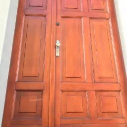 Ciemnobrązowe, drewniane drzwi wejściowe z metalową klamką i szybą u góry, widziane z dołu.