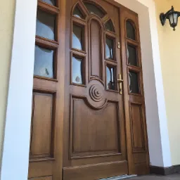 Eleganckie, przeszklone drzwi wejściowe z drewna w Gdyni, z dekoracyjnymi elementami i złotą klamką, oświetlone przez zewnętrzną lampę.