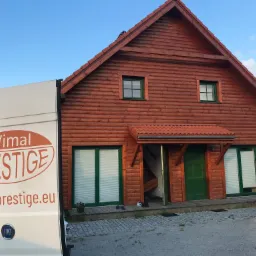 Drewniany dom z zielonymi oknami i drzwiami, częściowo zasłonięty białym vanem z logo firmy Wimal Prestige; plisy widoczne w oknach.