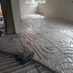 Rozłożone rury ogrzewania podłogowego na folii z siatką, widoczny worek z zaprawą i fragment narzędzia, surowe ściany w tle.