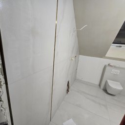 Remoncik Usługi Remontowo-Budowlane Marcin Piech - Remont łazienki na poddaszu: białe płytki z złotymi listwami, wisząca toaleta, okno dachowe. Częściowo wykończona ściana z widocznym murem.