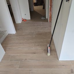Remoncik Usługi Remontowo-Budowlane Marcin Piech - Jasne, drewnopodobne płytki podłogowe w korytarzu po remoncie. Widoczna szczotka oparta o ścianę, surowe ściany i otwarte przejście do kolejnego pomieszczenia.