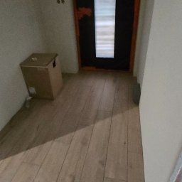 Remoncik Usługi Remontowo-Budowlane Marcin Piech - Wnętrze z drewnopodobną podłogą, ciemnymi drzwiami z przeszkleniem i kartonem przy ścianie. Widoczne ślady po wierceniu w ścianie.