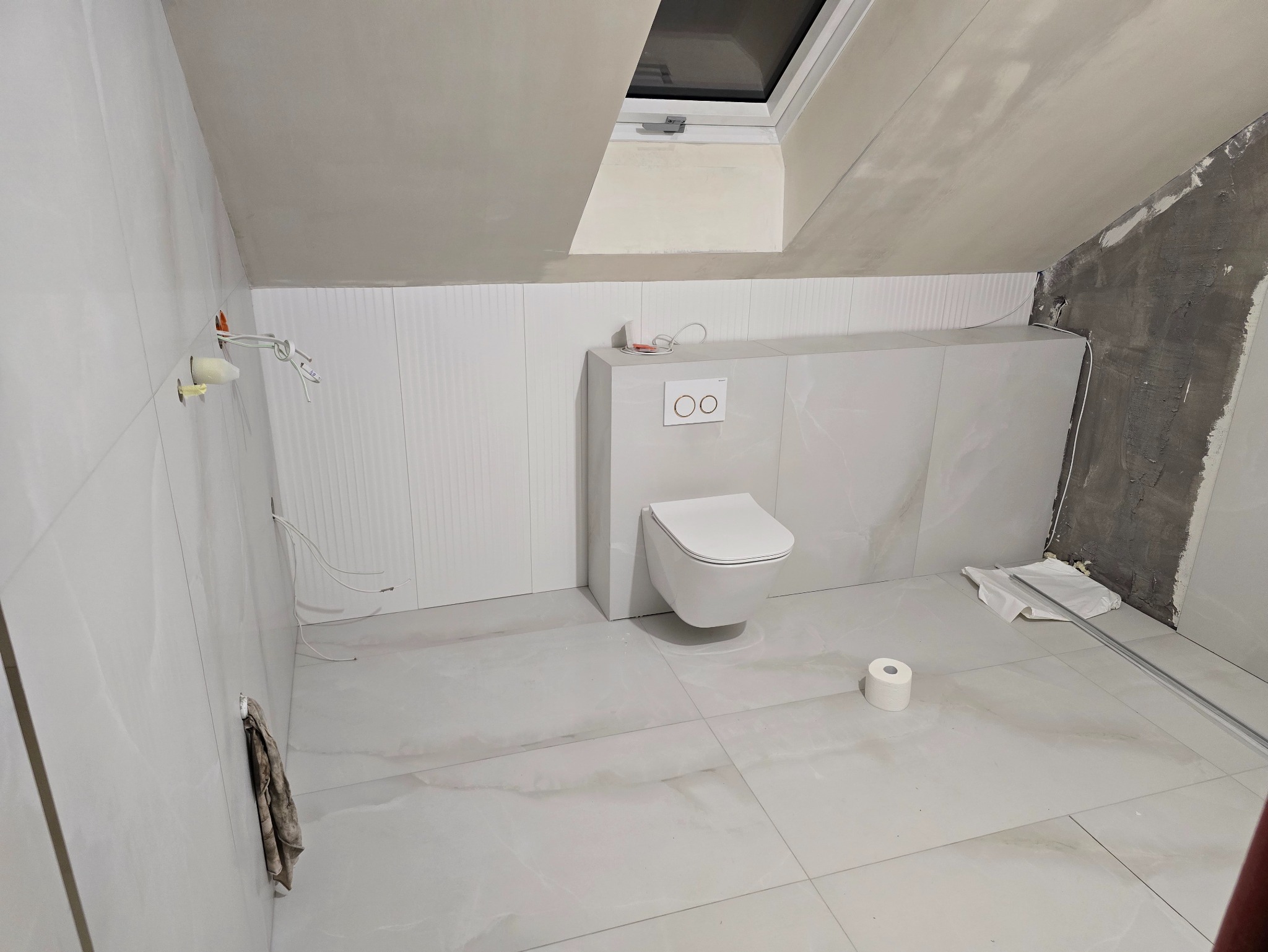 Wykończona łazienka na poddaszu ze skośnym sufitem i oknem dachowym. Białe płytki na ścianach i podłodze, wisząca toaleta. Częściowo widoczne przewody elektryczne.