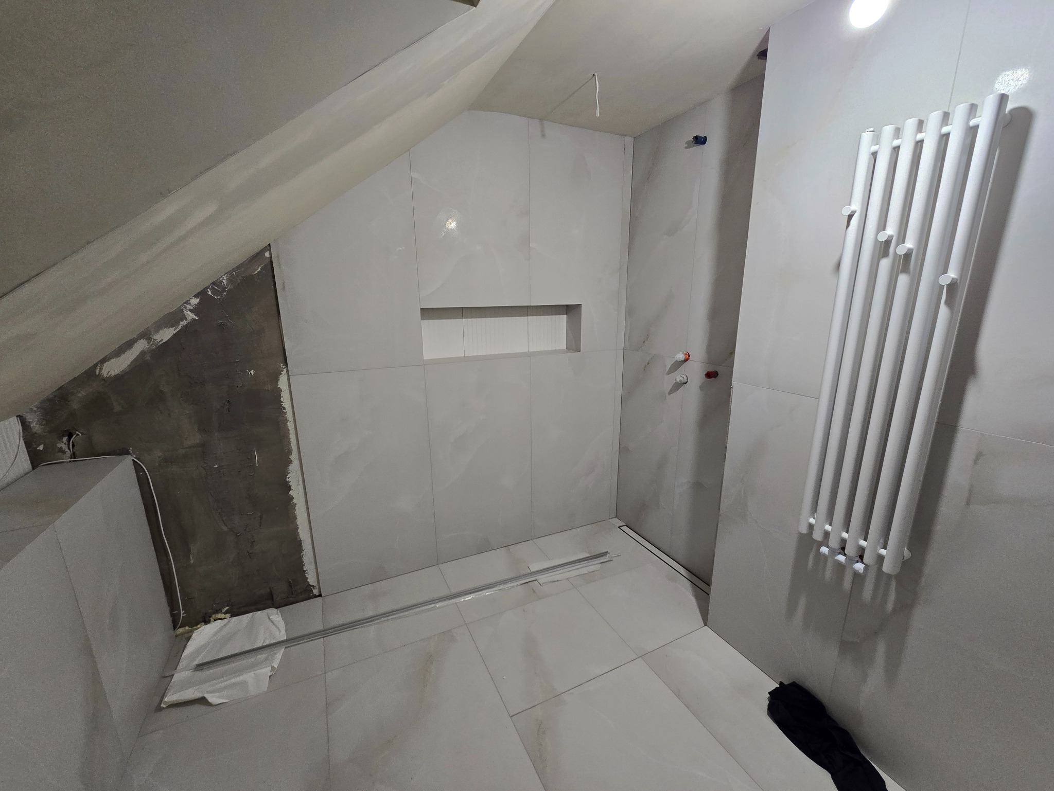 Łazienka na poddaszu w trakcie wykańczania: szare płytki, biały grzejnik drabinkowy, wnęka w ścianie. Część ściany surowa, przygotowana pod wykończenie.