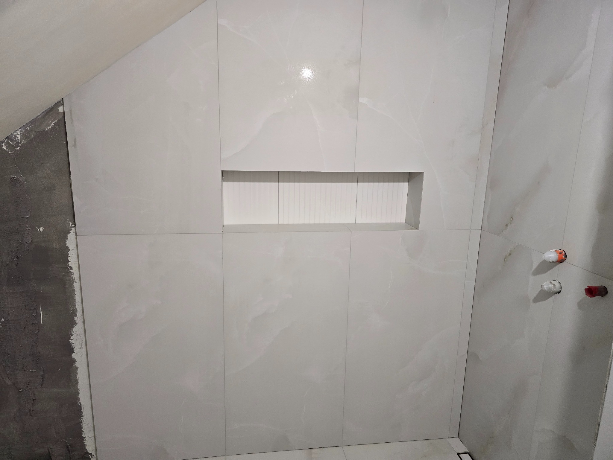 Wykończona ściana w łazience z jasnych, marmurkowych płytek. W ścianie wnęka z białymi, pionowymi panelami. Widoczne przyłącza wodne. Część ściany w trakcie prac.