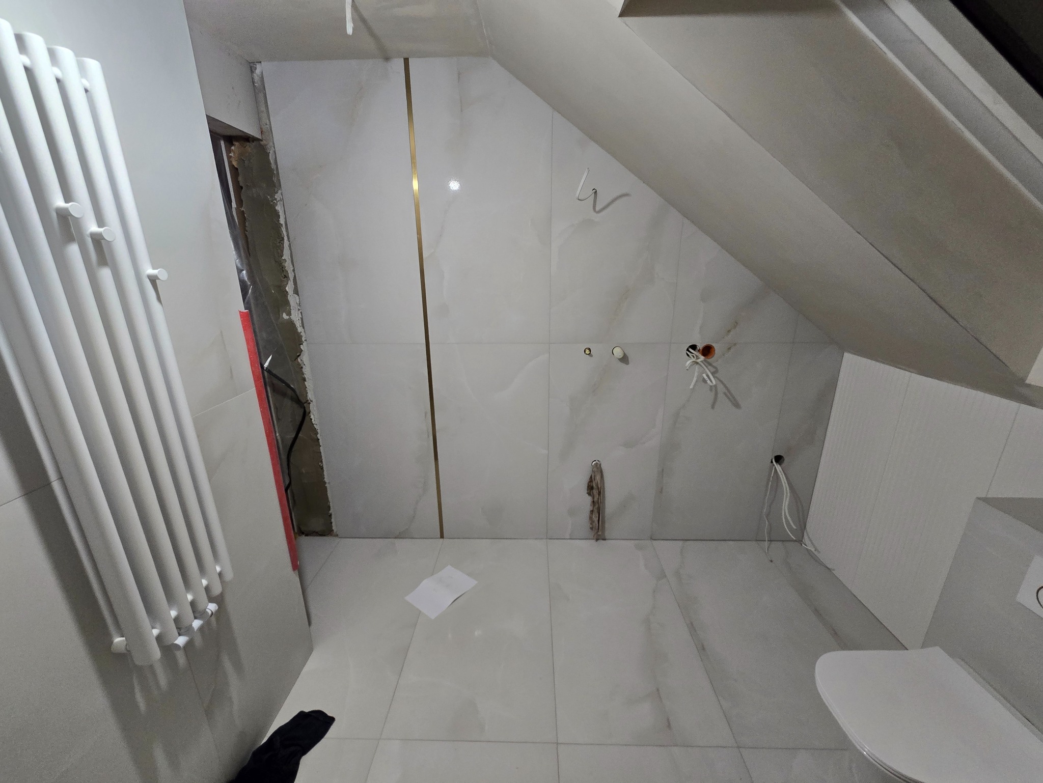 Wykończona łazienka na poddaszu ze skośnym sufitem. Białe płytki imitujące marmur z złotymi listwami, grzejnik drabinkowy i częściowo widoczna toaleta.