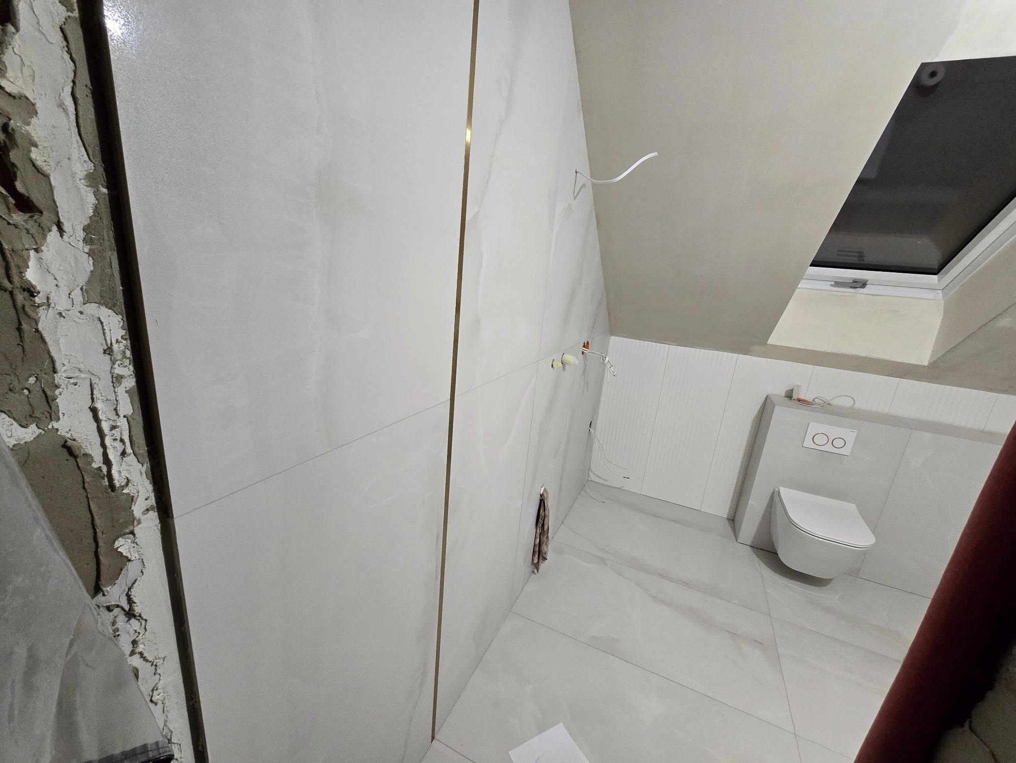 Remont łazienki na poddaszu: białe płytki z złotymi listwami, wisząca toaleta, okno dachowe. Częściowo wykończona ściana z widocznym murem.