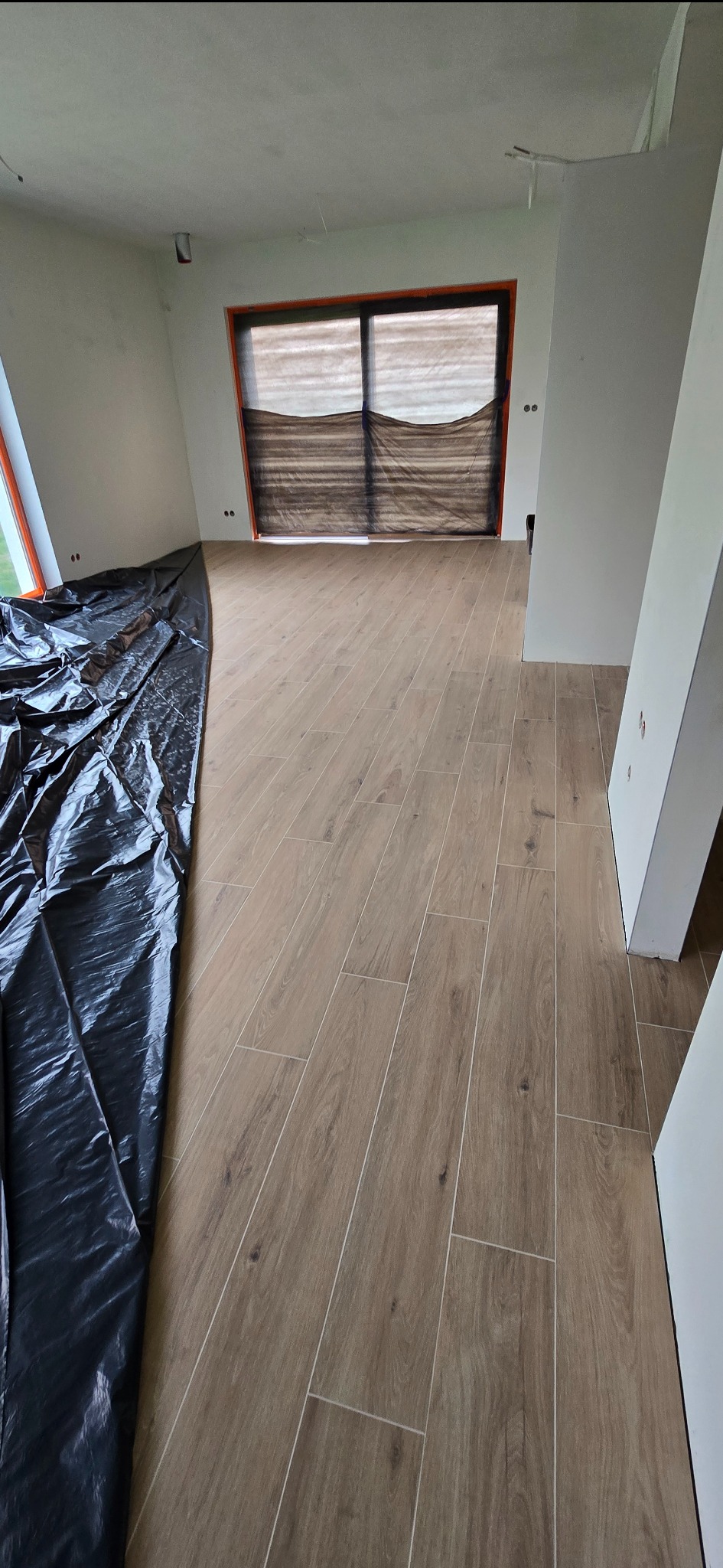 Ułożona podłoga z płytek imitujących drewno w jasnym wnętrzu. Część podłogi zabezpieczona czarną folią ochronną. Widoczne okno z pomarańczową ramą.