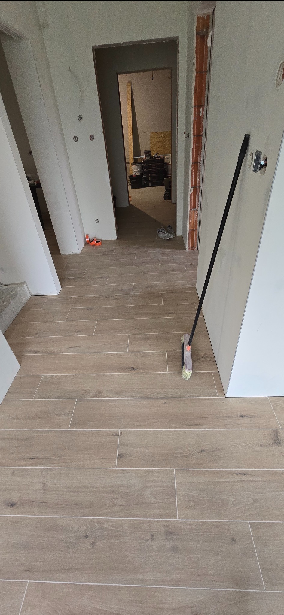 Jasne, drewnopodobne płytki podłogowe w korytarzu po remoncie. Widoczna szczotka oparta o ścianę, surowe ściany i otwarte przejście do kolejnego pomieszczenia.