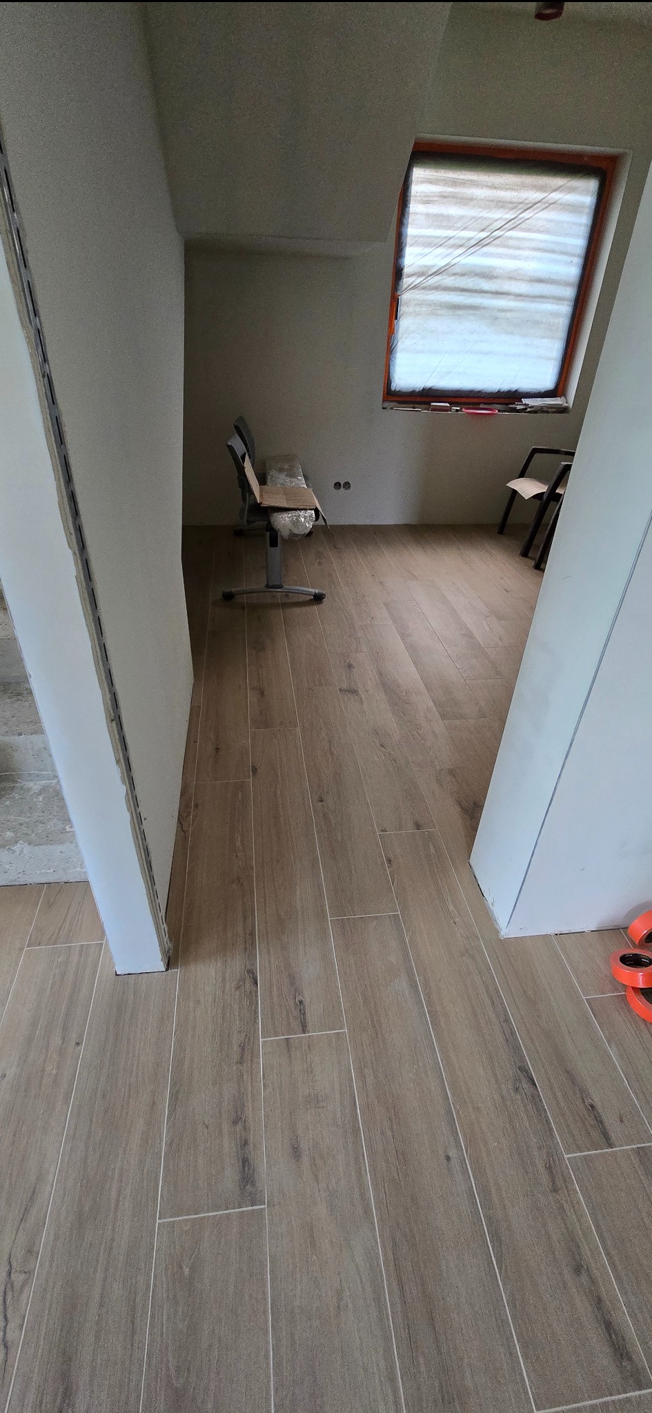 Ukończona podłoga z płytek imitujących drewno w jasnym pomieszczeniu z oknem dachowym. Widoczne krzesła i fragmenty ścian w trakcie wykończenia.