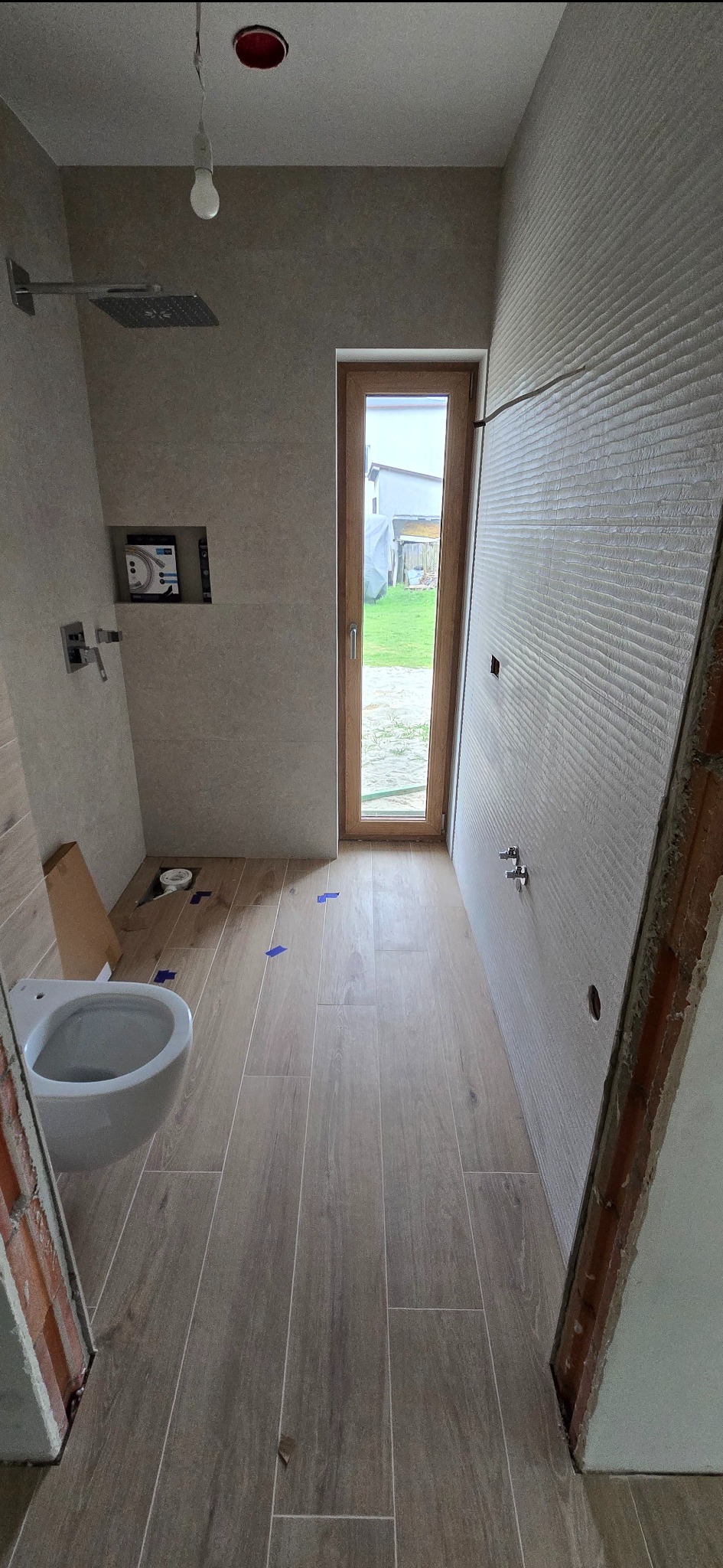 Wnętrze łazienki w trakcie wykańczania: ułożone płytki podłogowe imitujące drewno, częściowo wykończone ściany z widoczną instalacją, okno z widokiem na zieleń.