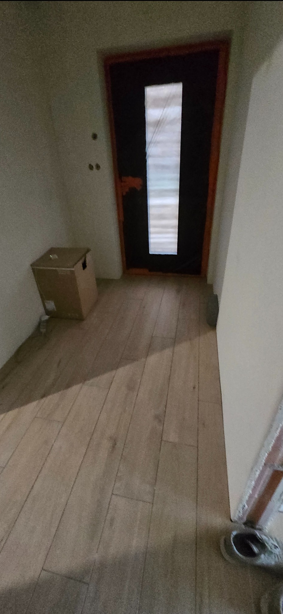 Wnętrze z drewnopodobną podłogą, ciemnymi drzwiami z przeszkleniem i kartonem przy ścianie. Widoczne ślady po wierceniu w ścianie.