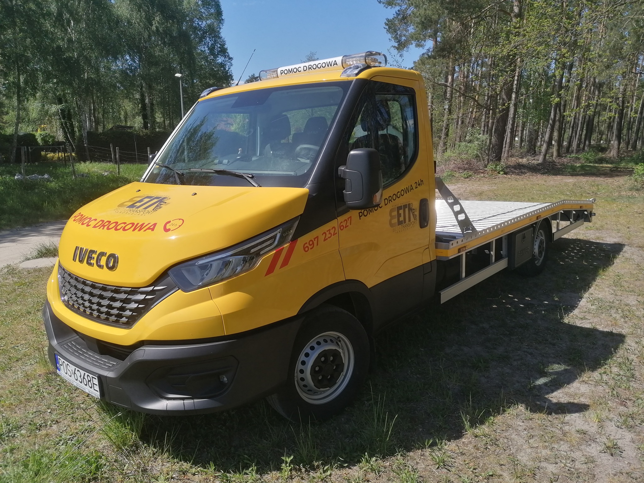 Żółta laweta Iveco z napisem 'Pomoc Drogowa' na tle zieleni, zaparkowana na trawie.