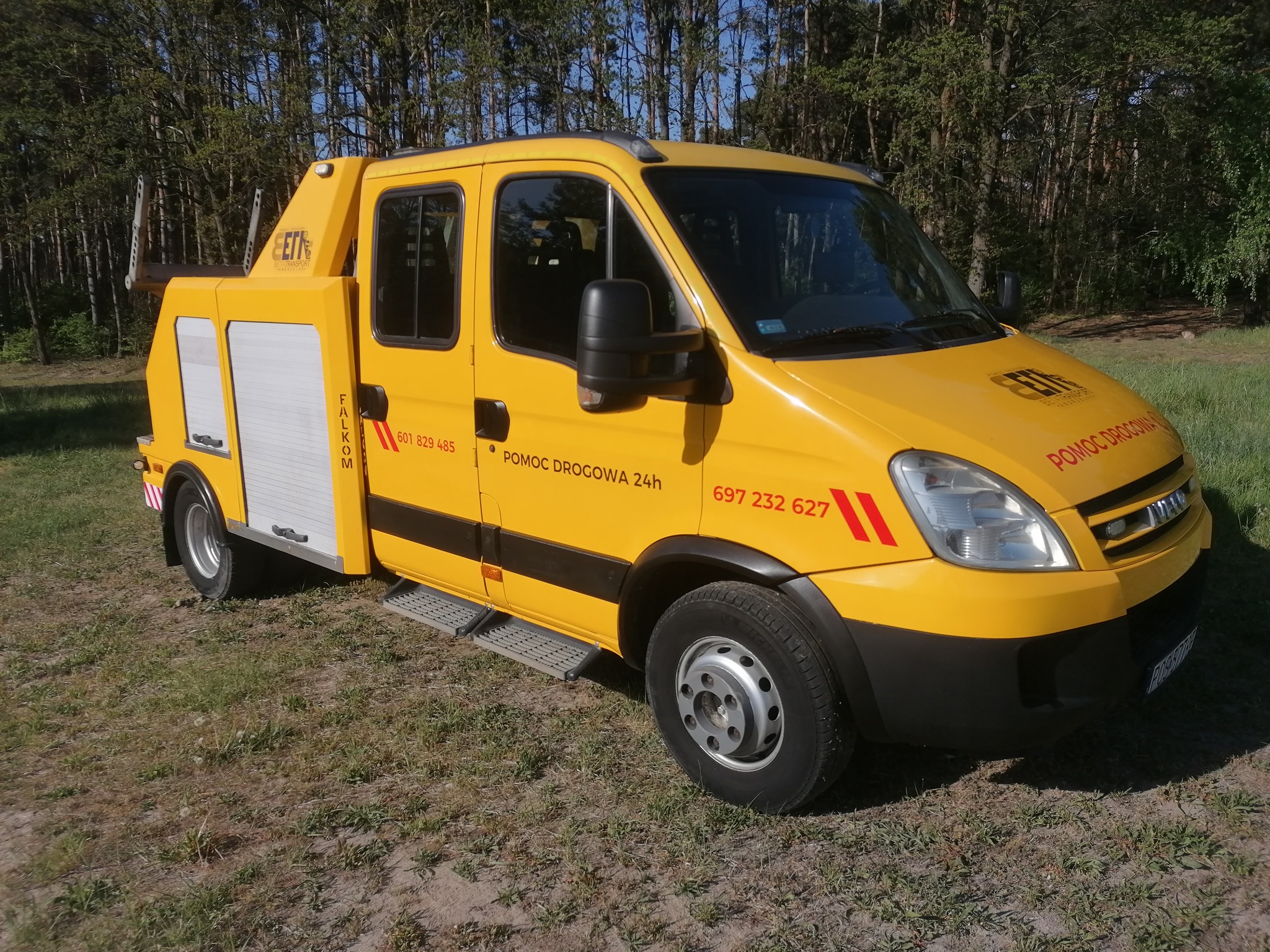 Żółty pojazd pomocy drogowej Iveco z napisem 'Pomoc Drogowa 24h' zaparkowany na trawiastym terenie, widoczne numery telefonów kontaktowych i logo firmy.