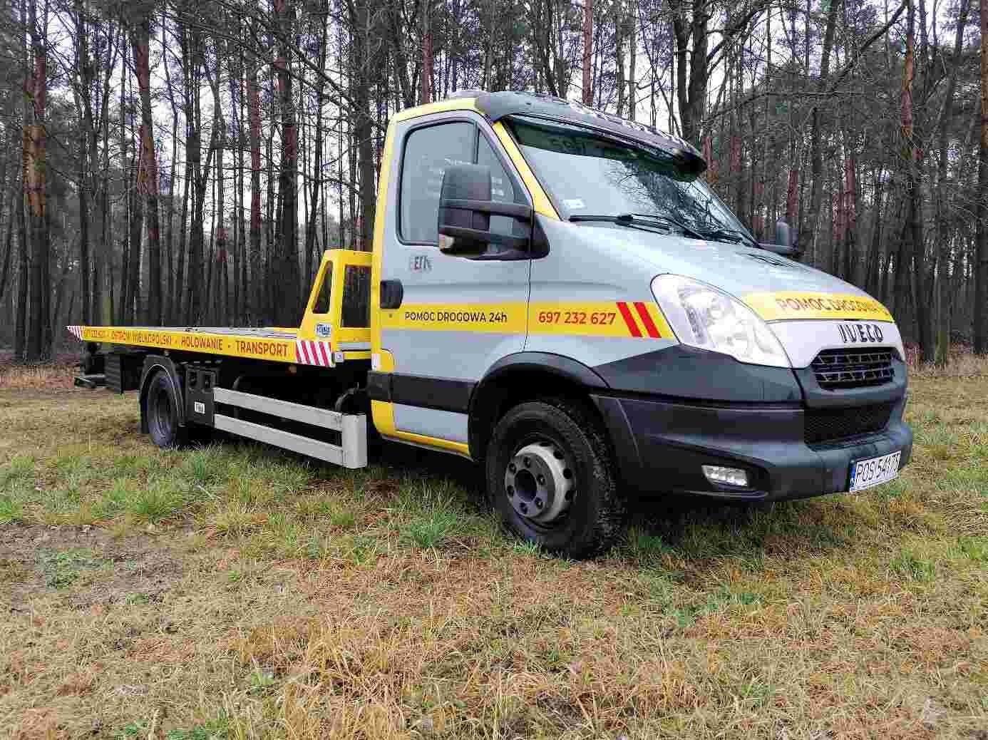 Szaro-żółta laweta Iveco z napisem 'Pomoc Drogowa 24h' zaparkowana na trawiastym terenie z widocznym lasem w tle. Na platformie lawety widoczne napisy informacyjne.