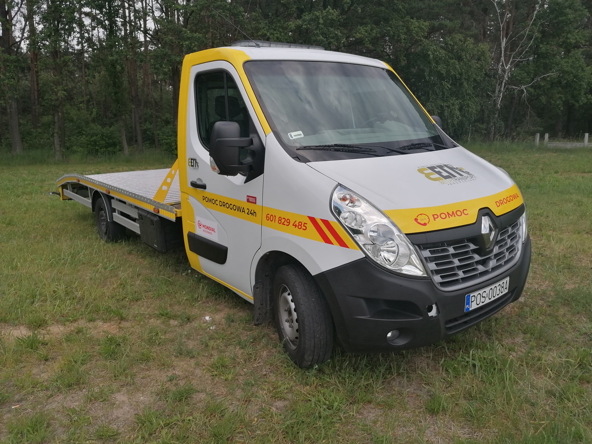 Biało-żółta laweta Renault Master z napisem 'Pomoc Drogowa 24h' i numerem telefonu, stojąca na trawiastym terenie.