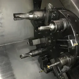 Głowica rewolwerowa tokarki CNC z zamocowanymi narzędziami skrawającymi, widoczne wiertła i noże tokarskie, wnętrze maszyny z widocznymi śladami chłodziwa.