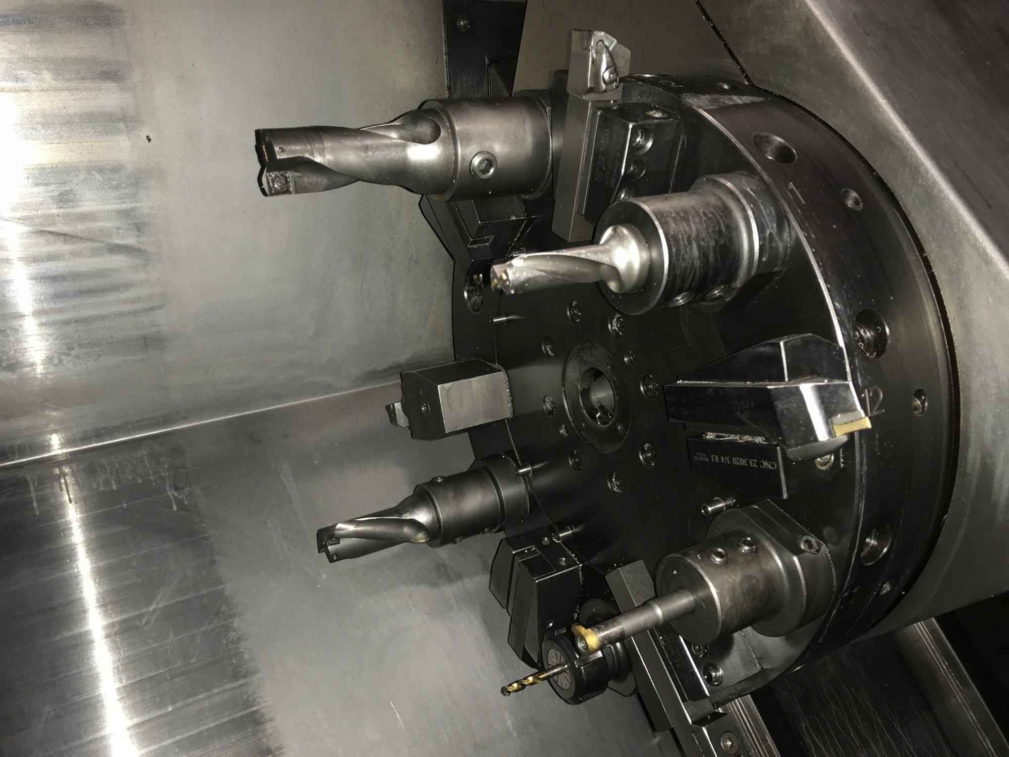 Głowica rewolwerowa tokarki CNC z zamocowanymi narzędziami skrawającymi, widoczne wiertła i noże tokarskie, wnętrze maszyny z widocznymi śladami chłodziwa.
