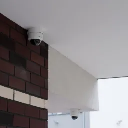 Dwie kamery kopułkowe monitoringu zamontowane na zewnątrz budynku z ceglaną ścianą i białym sufitem, widok z dołu.