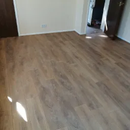 Ukończone układanie paneli podłogowych w jasnym wnętrzu z dwiema parami ciemnych drzwi, widoczna listwa przypodłogowa i gniazdko elektryczne na ścianie.