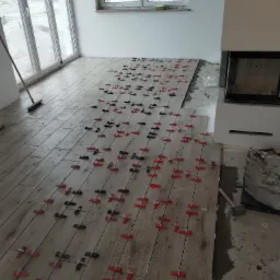 Układanie płytek podłogowych imitujących drewno w jasnym pomieszczeniu, z użyciem czerwonych i czarnych krzyżyków dystansowych, widok na częściowo ułożoną podłogę i przygotowane podłoże przy kominku.