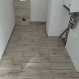 Ułożona podłoga z płytek imitujących drewno w łazience, widoczne przyłącza do toalety podtynkowej i piec gazowy, świeżo położone fugi.