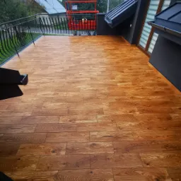 Balkon z wykończoną podłogą z desek w odcieniach brązu, z czarną balustradą i widokiem na otoczenie, w tle pomarańczowy podnośnik koszowy.