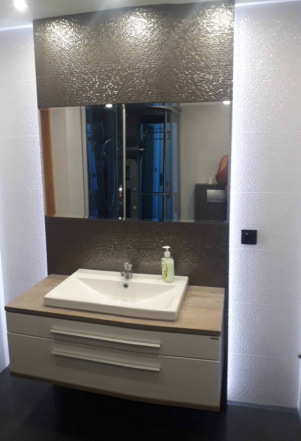 Nowoczesna łazienka z umywalką nablatową, szarą mozaiką nad umywalką i lustrem, oraz kabiną prysznicową widoczną w odbiciu lustra.