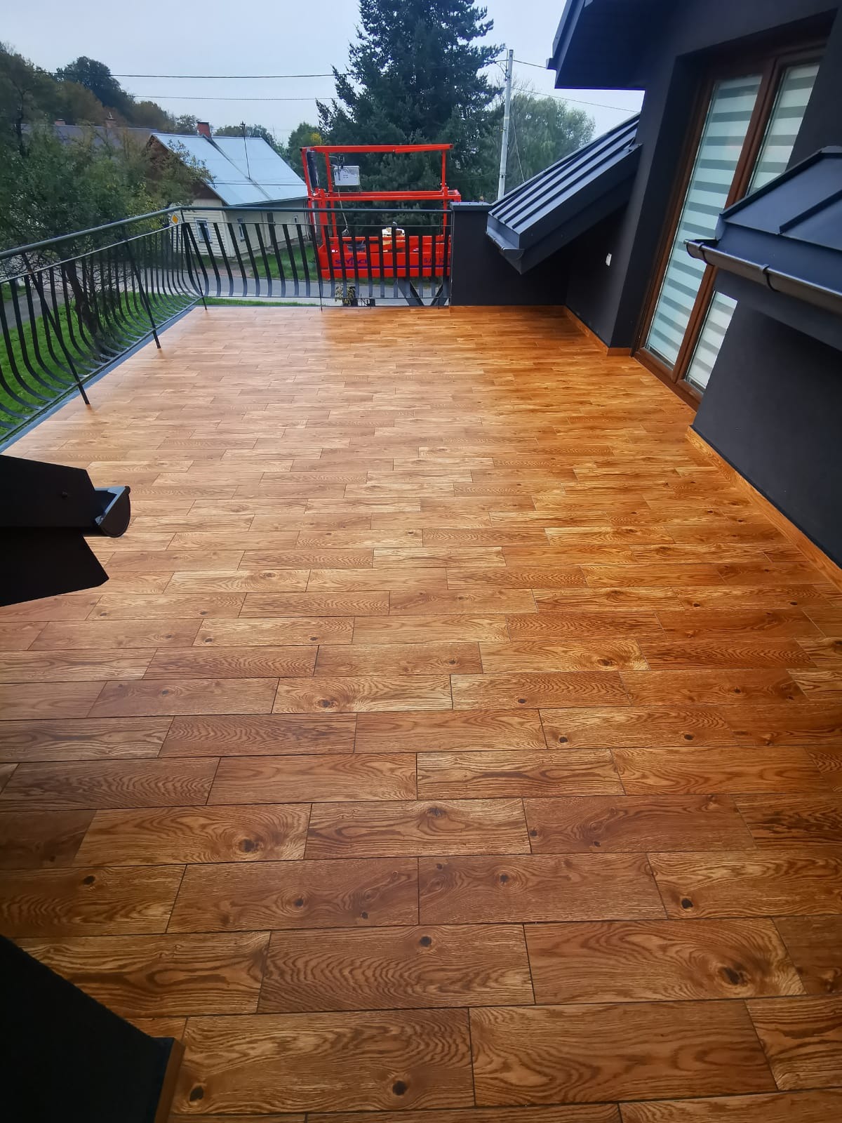 Balkon z wykończoną podłogą z desek w odcieniach brązu, z czarną balustradą i widokiem na otoczenie, w tle pomarańczowy podnośnik koszowy.